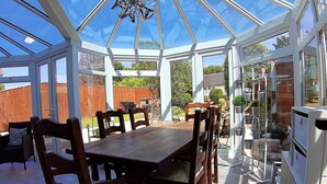 A conservatory with a wooden dining table and chairs, surrounded by glass walls and a view of the garden. - Charming Villa in Ryde - Child Friendly (Ryde)
