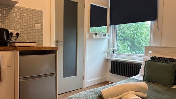 A compact hotel room with a kitchenette, a bed with a green blanket, a radiator, and a window with a view of greenery. - compact studio in central location with excellent transport links. (London)
