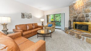 A living room with a stone fireplace, leather sofas, and a view of greenery through a door. - Holly Ridge 204 | Near Gatlinburg + Cozy (Gatlinburg)