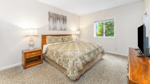A bedroom with a bed, two bedside tables with lamps, a TV stand, a window with blinds, and a painting on the wall. - Holly Ridge 204 | Near Gatlinburg + Cozy (Gatlinburg)