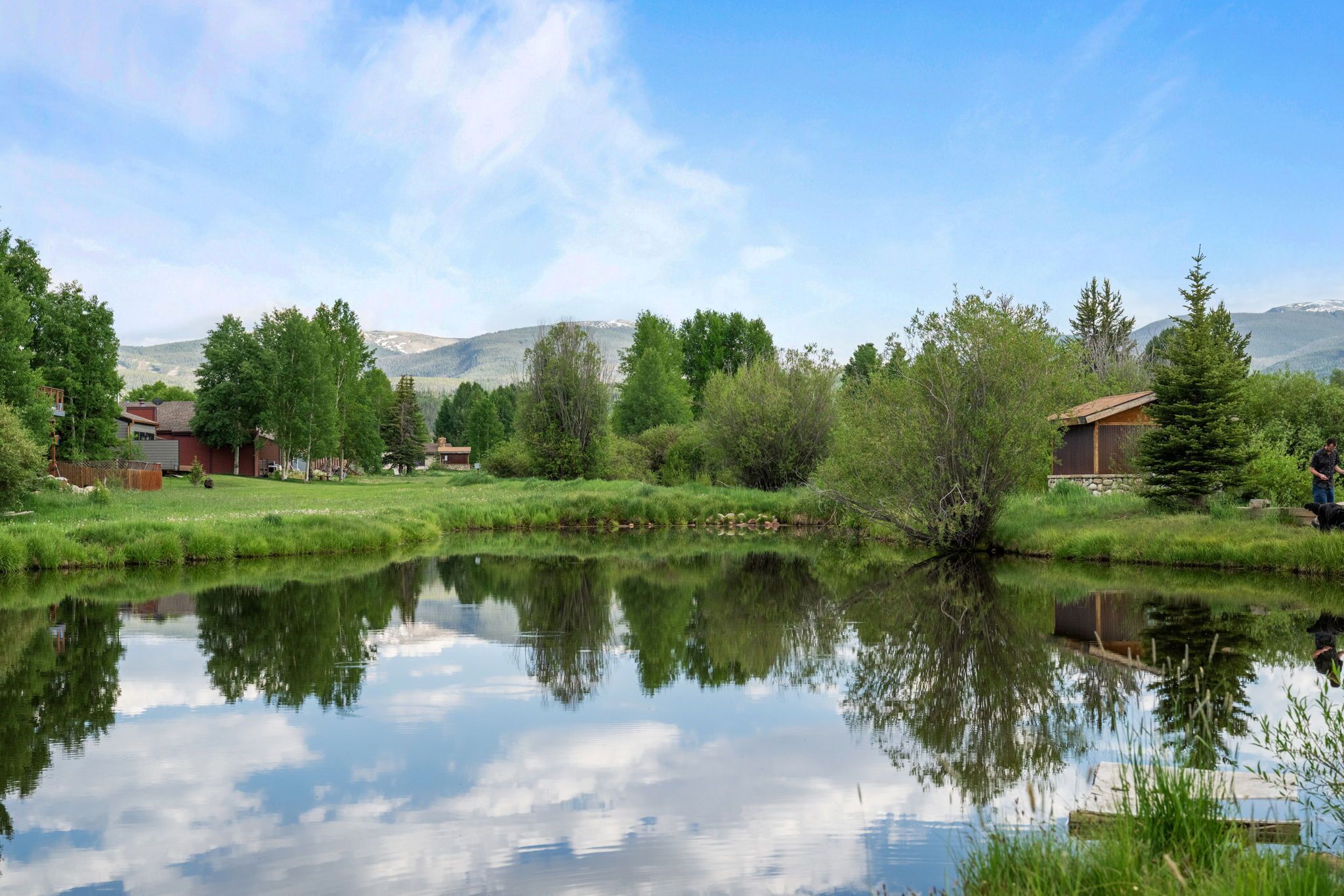 A serene lakeside view with a clear sky, reflecting trees and buildings, and a distant mountain range.