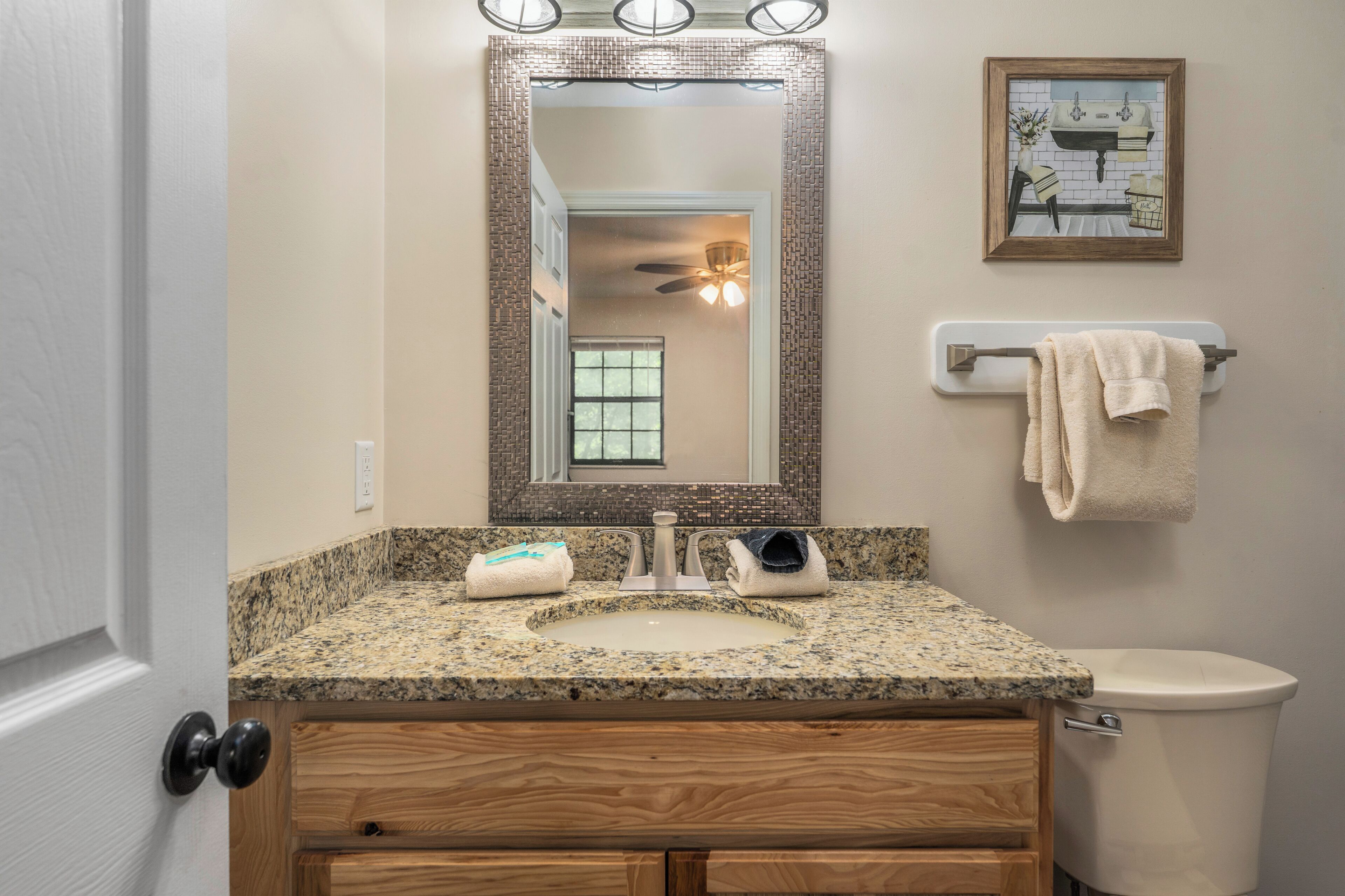 A bathroom with a granite countertop sink, wooden cabinetry, a toilet, a towel rack, and a mirror with a decorative frame.