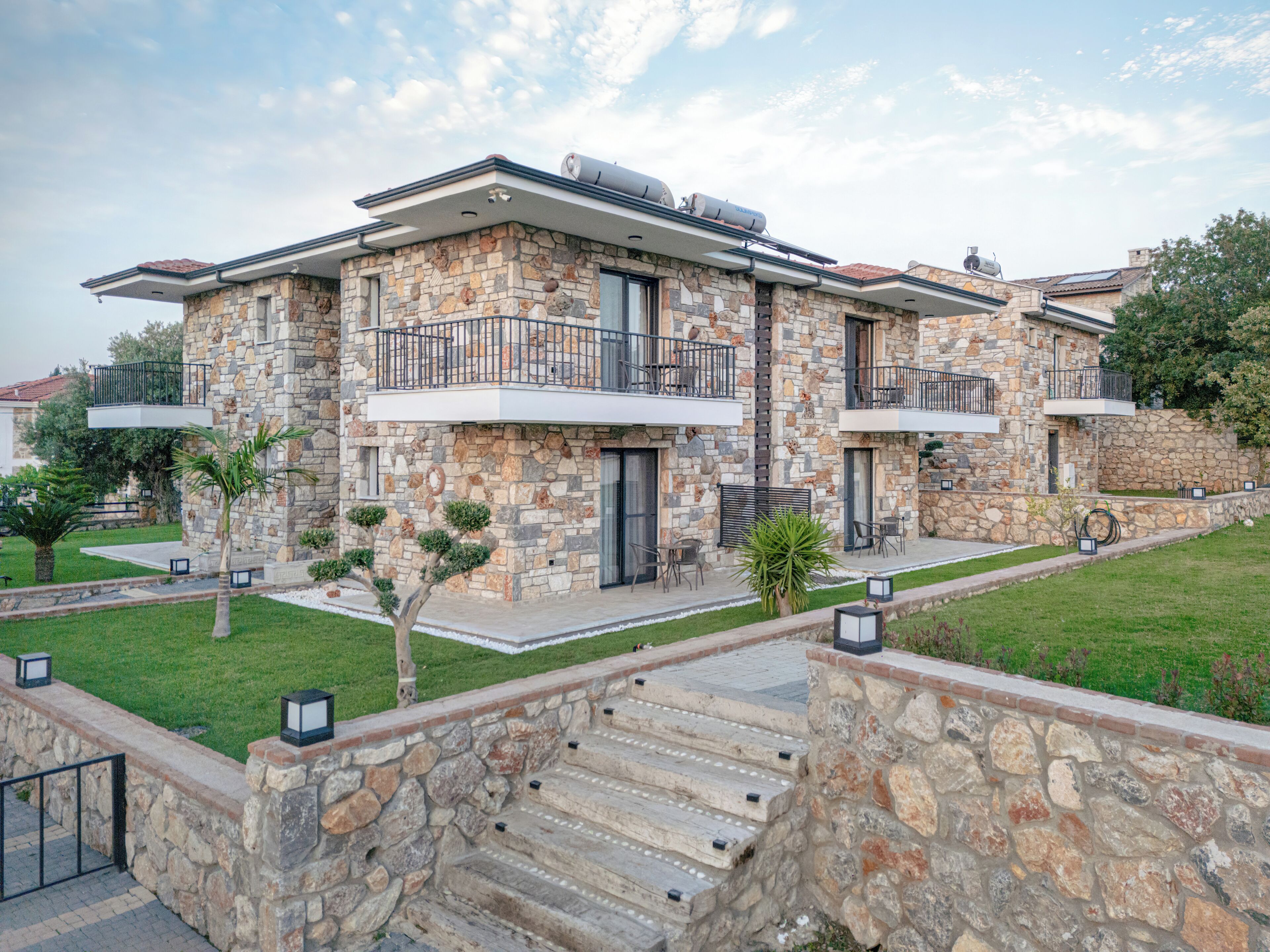 Une maison moderne à deux étages, dotée d’une façade en pierre, de balcons et d’un jardin bien entretenu.