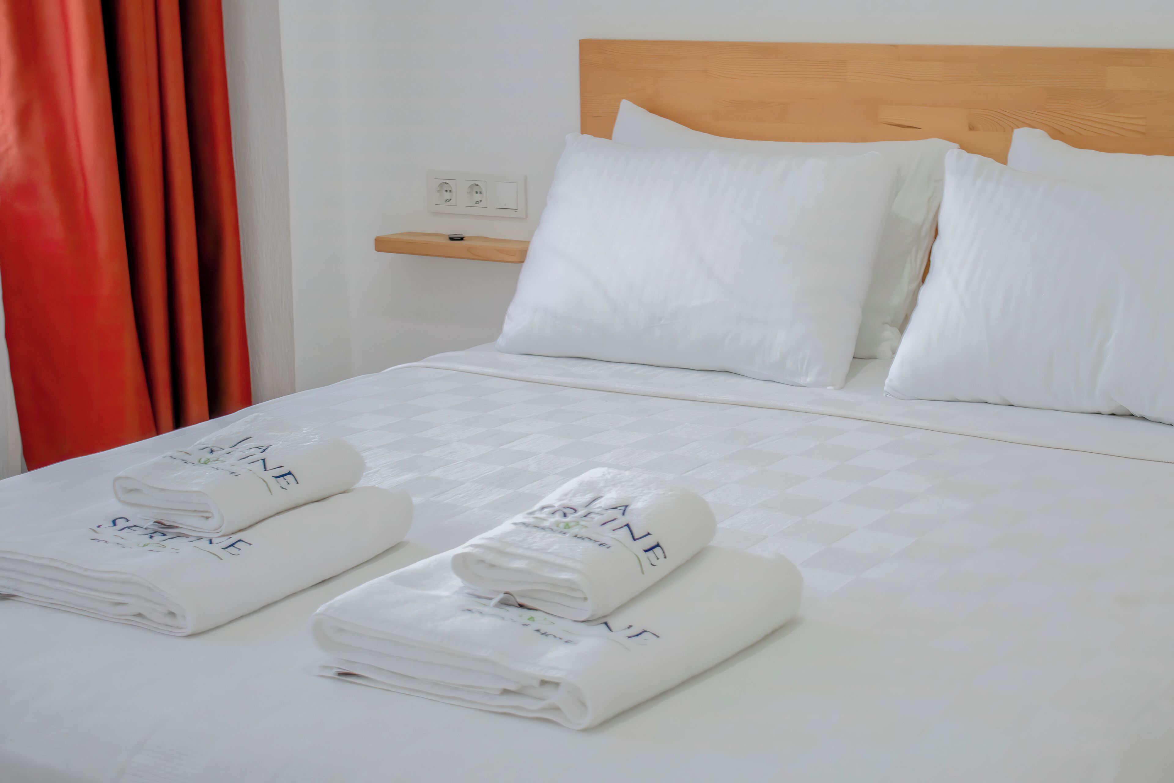 A neatly made bed with white linens and towels, a wooden headboard, and a small shelf with a power outlet.