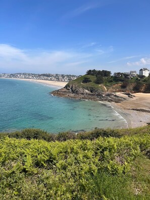 A coastal landscape with a beach, rocky outcrops, and a building on a hill.