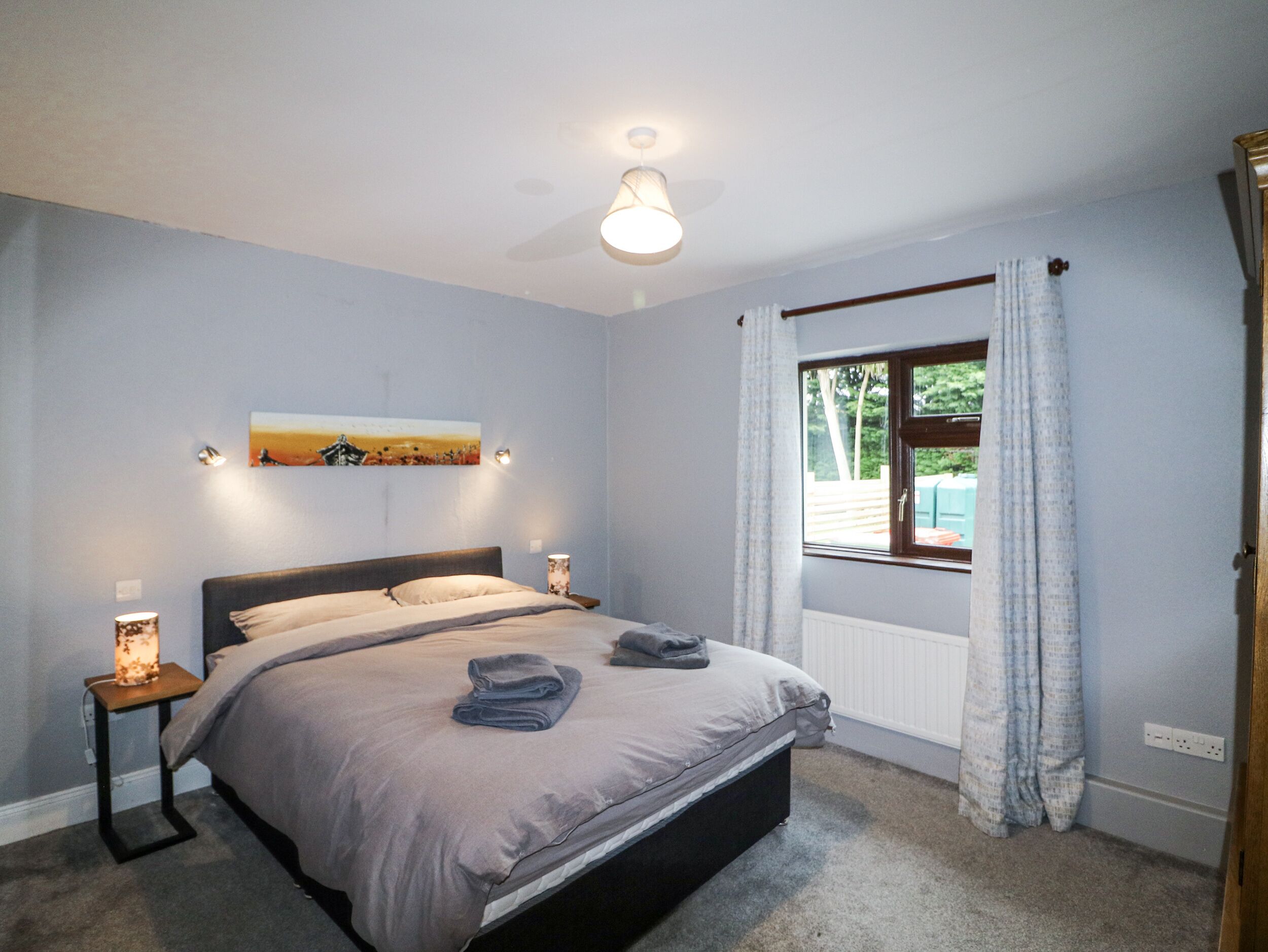 A bedroom with a bed, bedside lamps, a painting on the wall, a window with curtains, and a radiator.