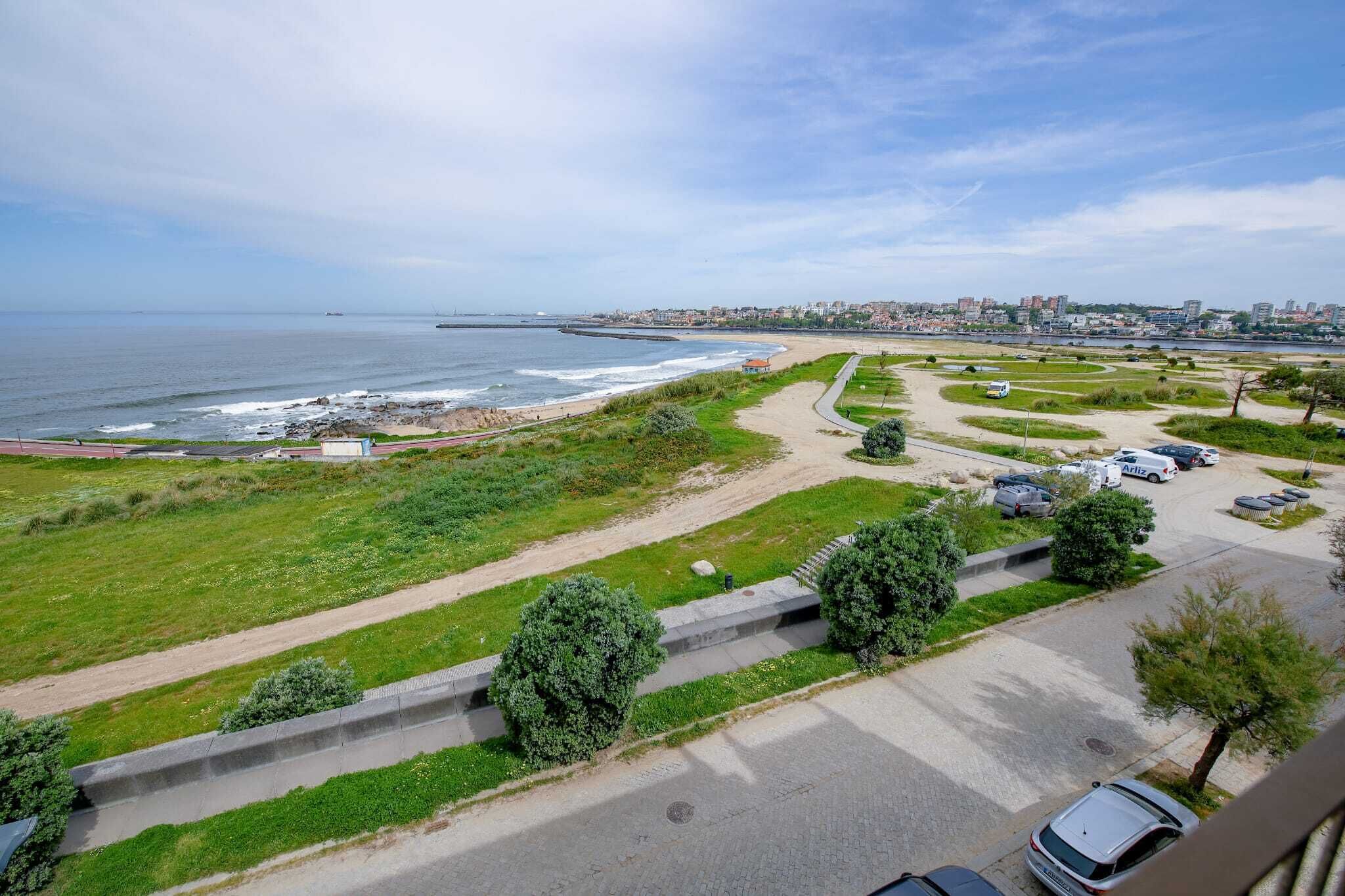 Vista costeira com praia, área de estacionamento e paisagem urbana ao fundo.