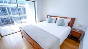 A bedroom with a wooden bed, white bedding, a nightstand with a lamp, and a view of a building through a large window. - Atlantis Ocean front (Vila Nova de Gaia)