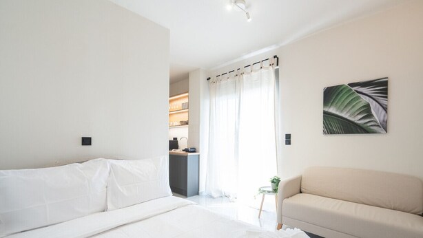 Une chambre d’hôtel avec un lit, un canapé, une petite table avec une plante, et un coin cuisine visible à travers une porte.