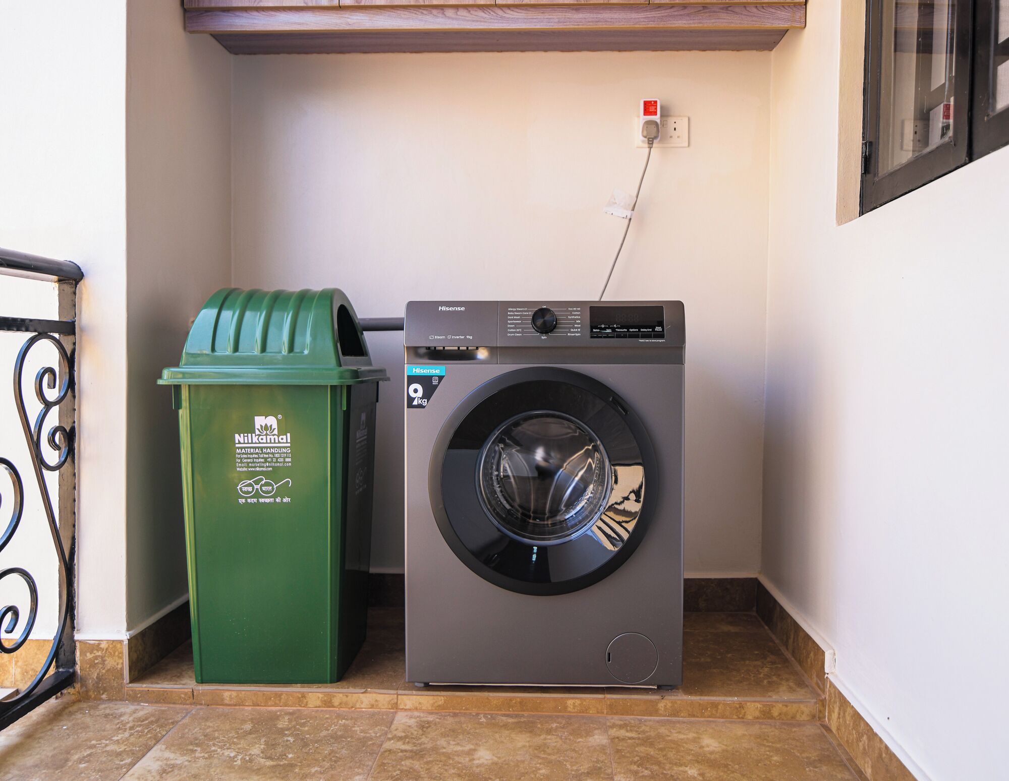 A compact washing machine and a green waste bin in a small laundry area with tiled flooring and a wooden shelf above.