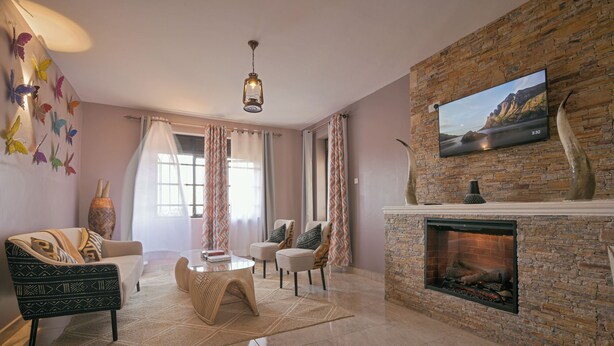 A living room with a fireplace, a television mounted on a brick wall, and a large window with curtains. - Aqueduct 1359 Hotel Apartments (Kampala)
