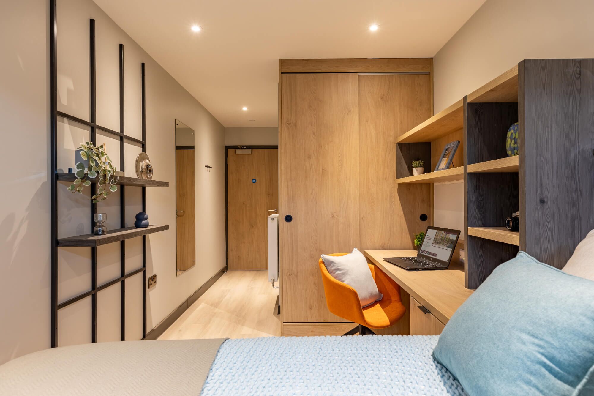 A modern bedroom with a bed, a desk with a laptop, a chair, and shelves with decorative items.