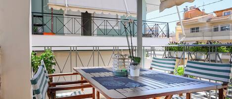 Un patio exterior con una mesa de madera y sillas a rayas, una planta en maceta y vistas a un edificio con balcones.