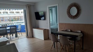 Habitación de hotel con balcón, televisión montada en la pared, zona de comedor con mesa y sillas, y pared revestida de paneles de madera.