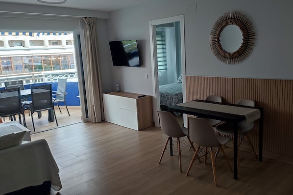 Habitación de hotel con balcón, televisión montada en la pared, zona de comedor con mesa y sillas, y pared revestida de paneles de madera.