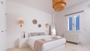A bedroom with a bed, bedside lamps, a mirror, and a window with blinds. - Golden Sunrise Suites - Santorini Lux Family Suite (Santorini)