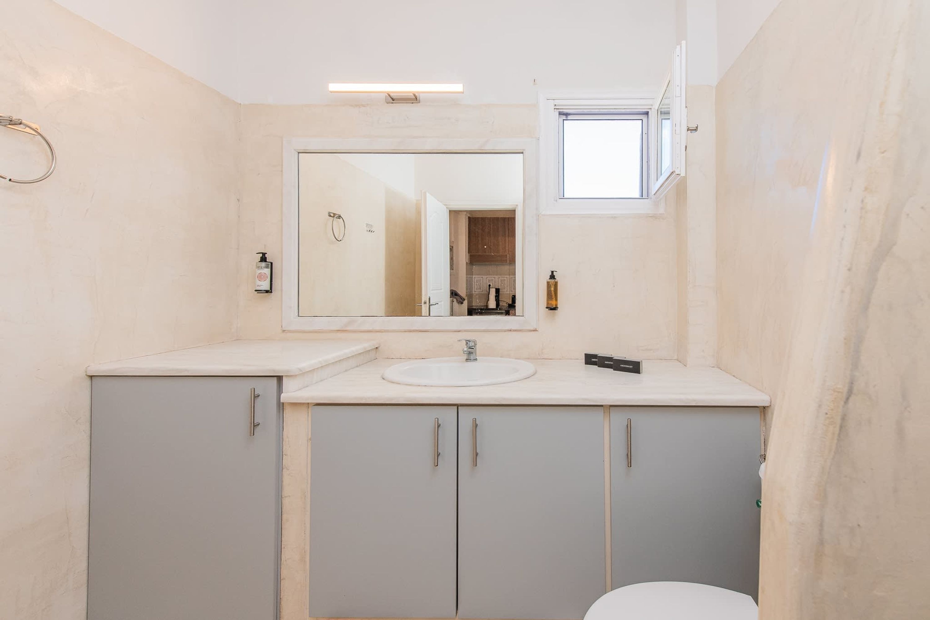 Ein Badezimmer mit einem weißen Waschbecken, einem Spiegel und einer Toilette. Über dem Spiegel befindet sich ein kleines Fenster.