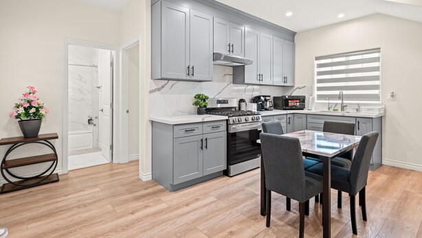 A modern kitchen with grey cabinets, a built-in oven, and a dining area with a table and chairs. - Modern Getaway Near Pasadena Dtla (Rosemead)