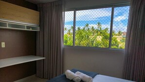 Une chambre d’hôtel avec un lit, une fenêtre donnant sur des palmiers et l’océan, et une étagère au-dessus du lit.