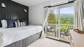 A bedroom with a bed, bedside lamps, a balcony with chairs, and a view of greenery.