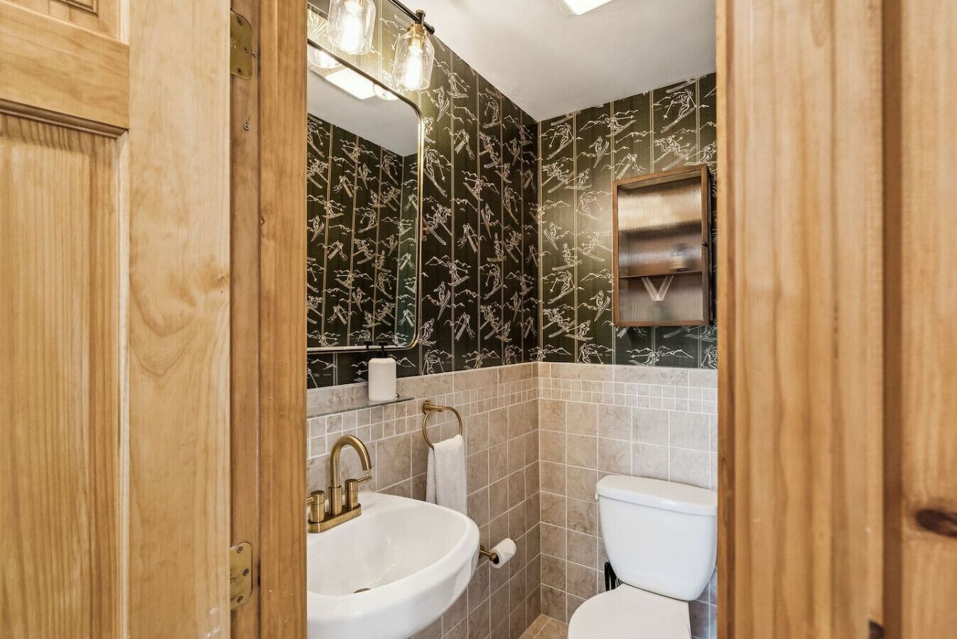 A bathroom with a wooden door, a white sink with gold fixtures, a toilet, and a mirror with a light fixture.
