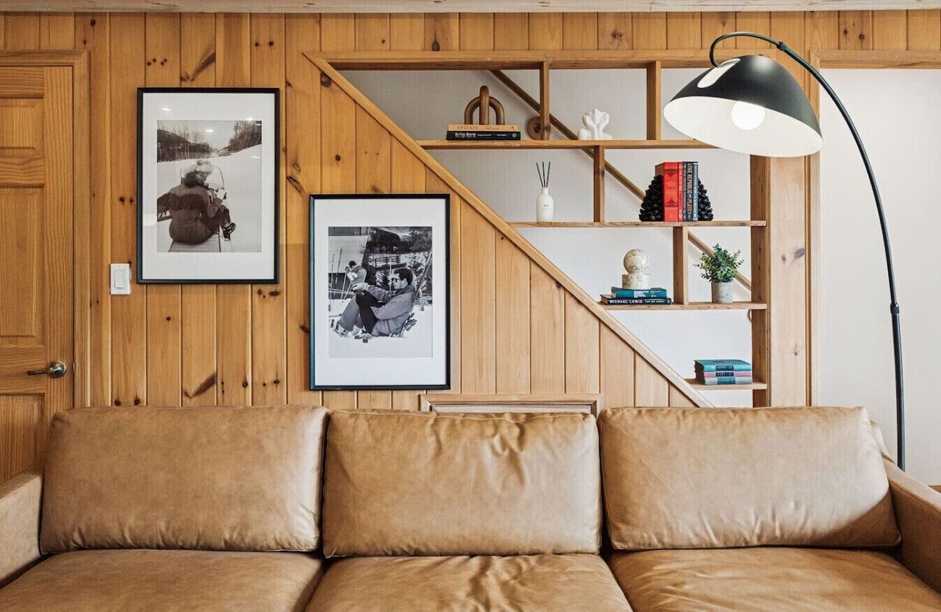 Ein Zimmer mit Holzwänden, einer Couch, zwei gerahmten Fotografien, einem Bücherregal und einer Stehlampe.