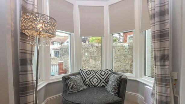 A corner room with a round grey sofa, patterned cushions, a decorative light fixture, and large windows with curtains. - Ty Llwyd (Llandudno)