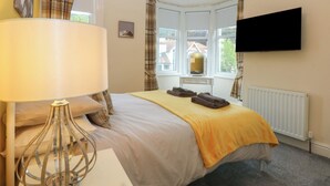 A bedroom with a bed, a bedside lamp, a dresser, a television, and a window with curtains. - Ty Llwyd (Llandudno)