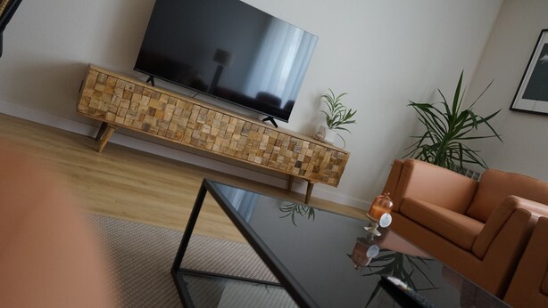 A modern living room with a flat-screen TV, a woven wooden console table, a glass coffee table, a leather sofa, and a potted plant. - Serendipity (Zaragoza)