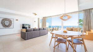 Una sala moderna con un sofá gris, una mesa de comedor redonda de madera y un gran ventanal con vistas al océano.