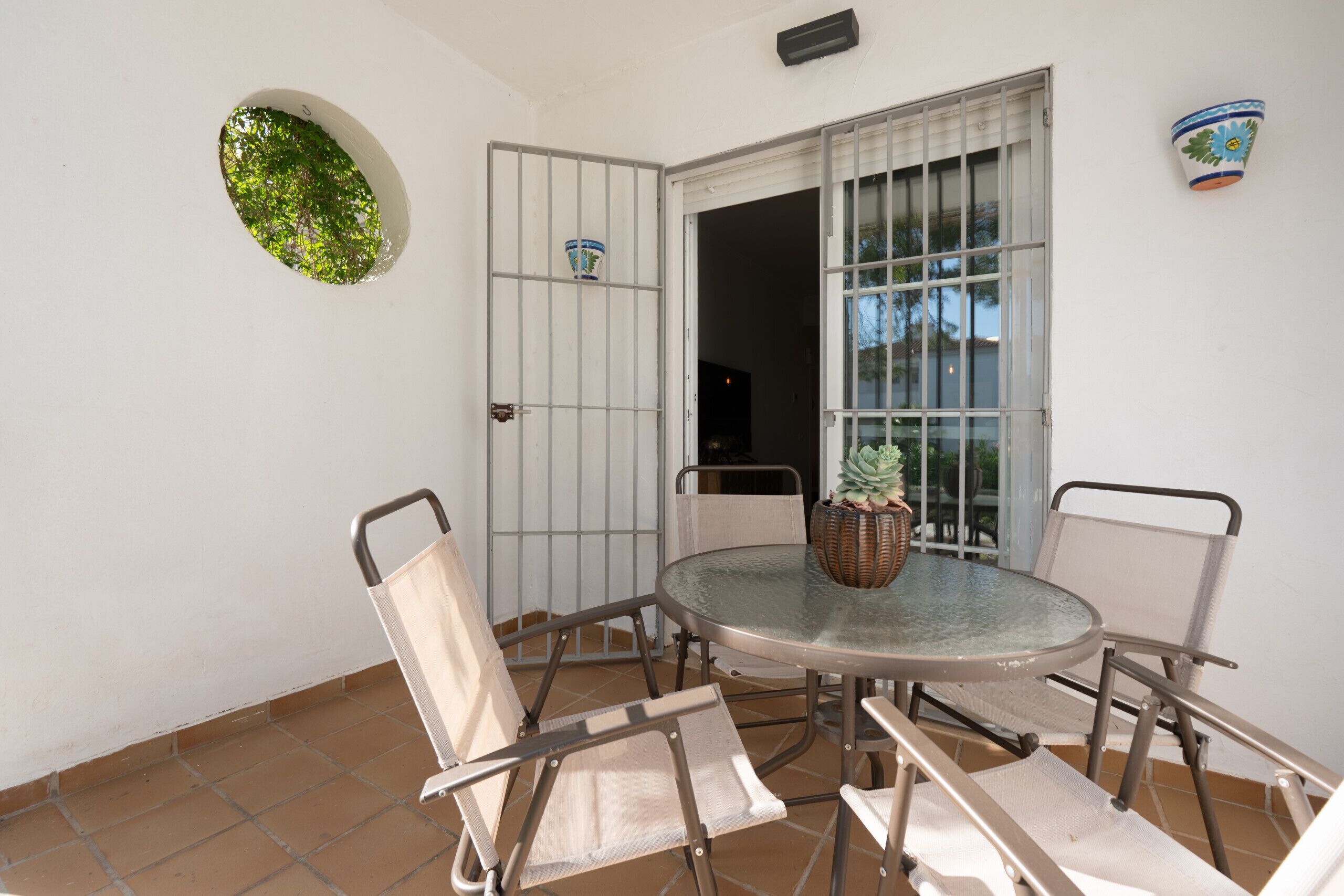 Une petite terrasse avec une table ronde en verre, quatre chaises en métal, un pot de fleurs et une fenêtre à barreaux.