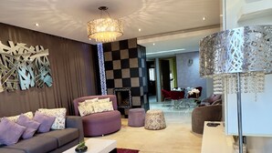 Un salon moderne avec un canapé gris, une table basse blanche et un tapis rouge. On y trouve une cheminée, un coin repas à l’arrière-plan et un élément décoratif mural.