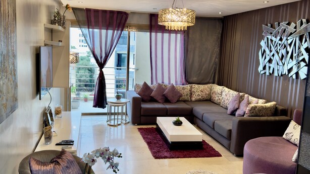 Un salon moderne avec un canapé d’angle, une table basse en verre et un pouf rond. On y trouve une petite table d’appoint avec un vase et une plante. La pièce est dotée d’une grande fenêtre avec des rideaux et offre une vue sur l’extérieur.