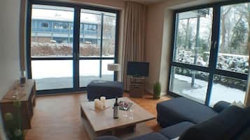Uma sala de estar moderna com um sofá, uma mesa de centro e uma televisão. Janelas amplas oferecem vista para o exterior.