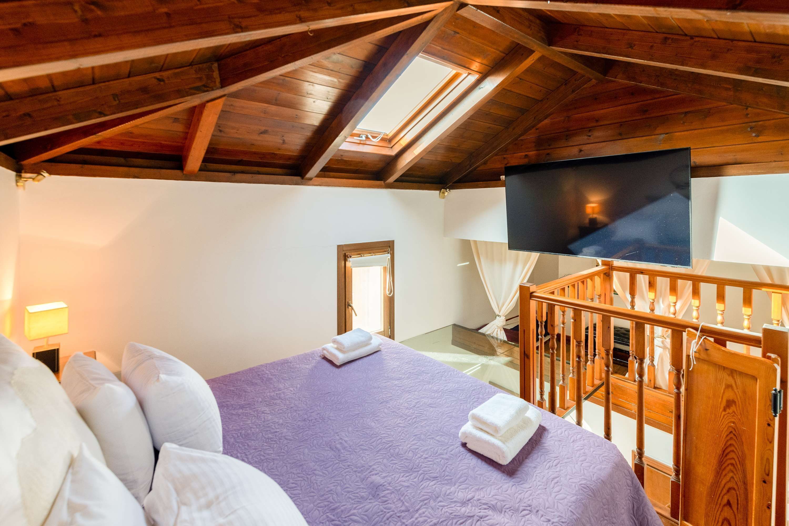 Une chambre avec un plafond en bois, un lit avec une literie blanche et violette, une télévision et un balcon doté d’une balustrade en bois.