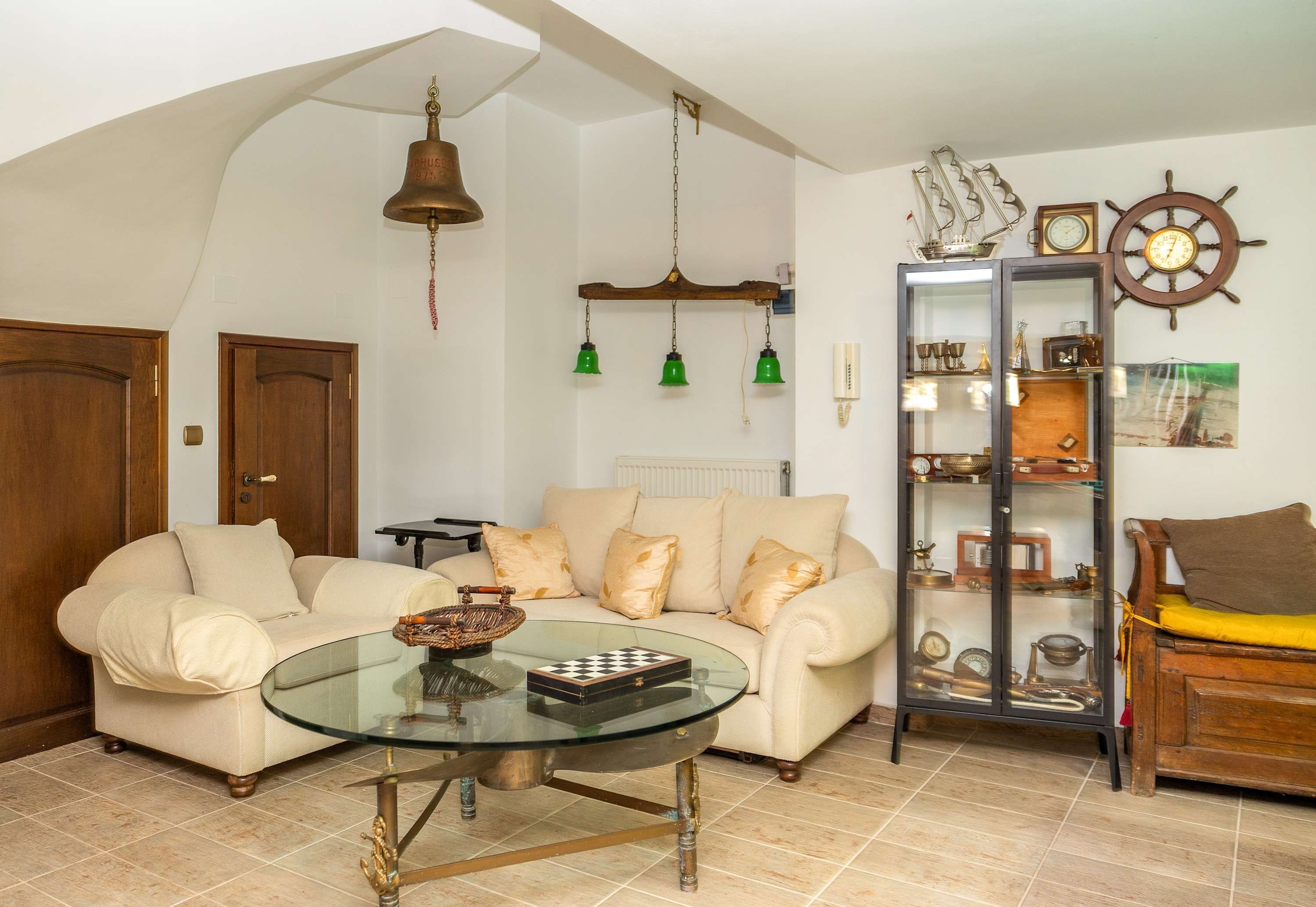A living room with a sofa, a glass coffee table, a wooden cabinet with glass doors, and a ship's wheel decoration on the wall.
