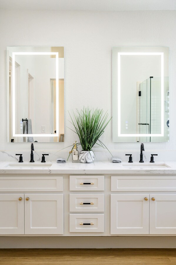 A bathroom with a white vanity, two mirrors with lights, a plant, and a marble countertop. - Luxe Spacious Home Near Parks Fully Equipped (Riverside)