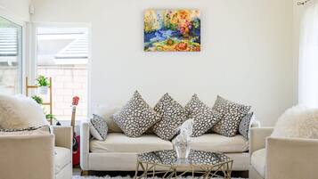 Un salon avec un canapé beige, des coussins à motifs, une table basse en verre et un tableau floral accroché au mur.