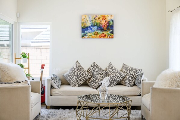 A living room with a beige sofa, patterned pillows, a glass coffee table, and a floral painting on the wall. - Luxe Spacious Home Near Parks Fully Equipped (Riverside)