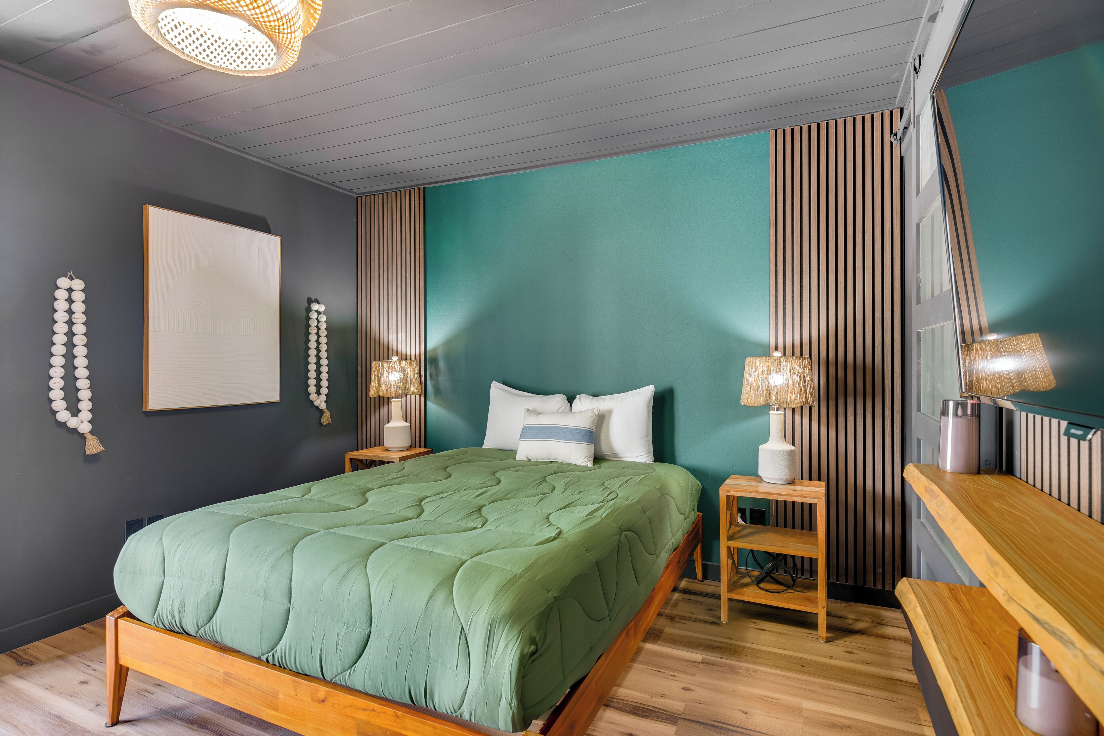 A bedroom with a green bed, wooden headboard, two bedside tables with lamps, a wall-mounted picture, and a bench.
