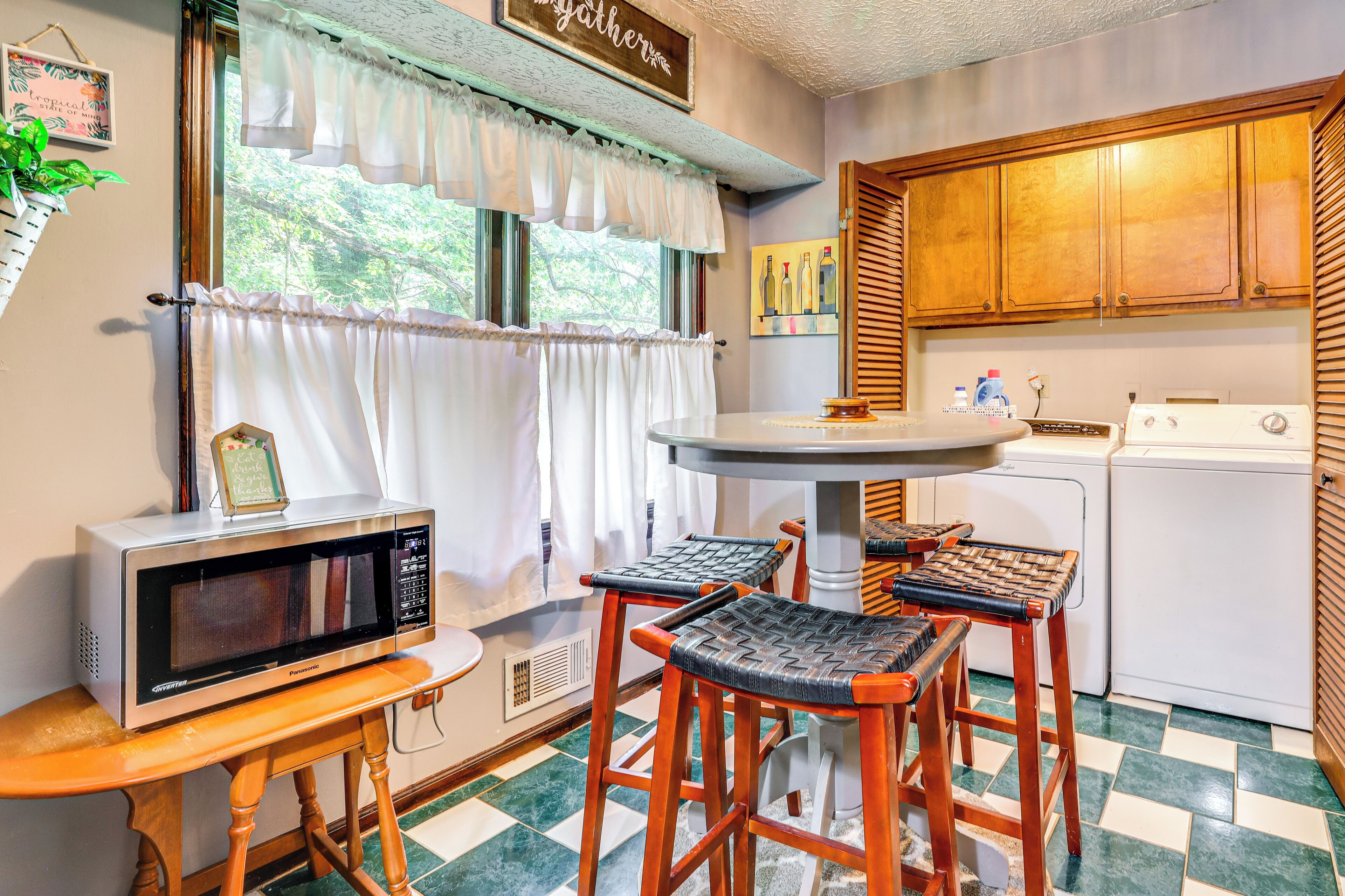 A kitchen with a microwave, a table, and two stools. There is a window with curtains, a washing machine, and wooden cabinets.