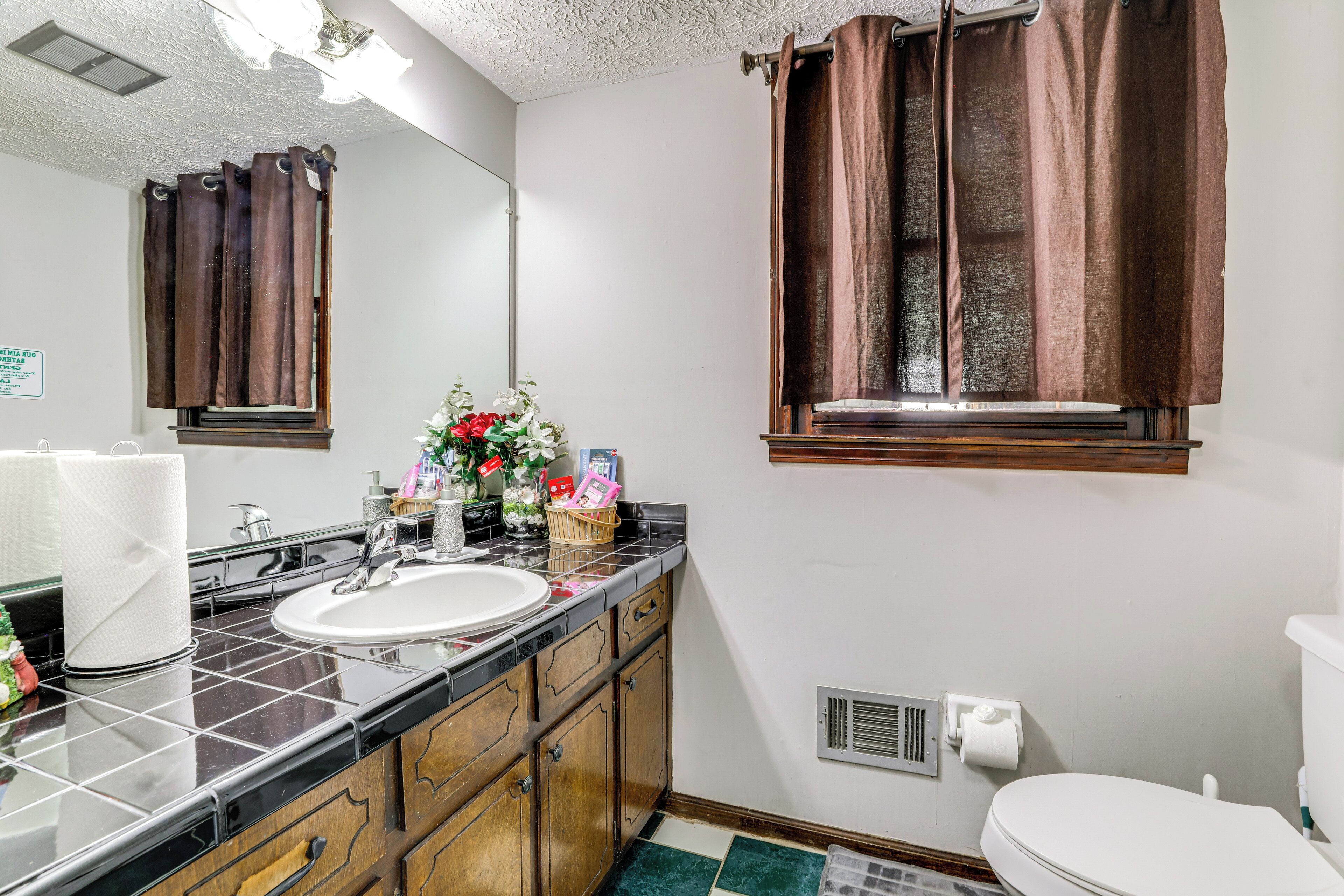 Un baño con lavamanos, espejo, cortinas marrones y una ventana.