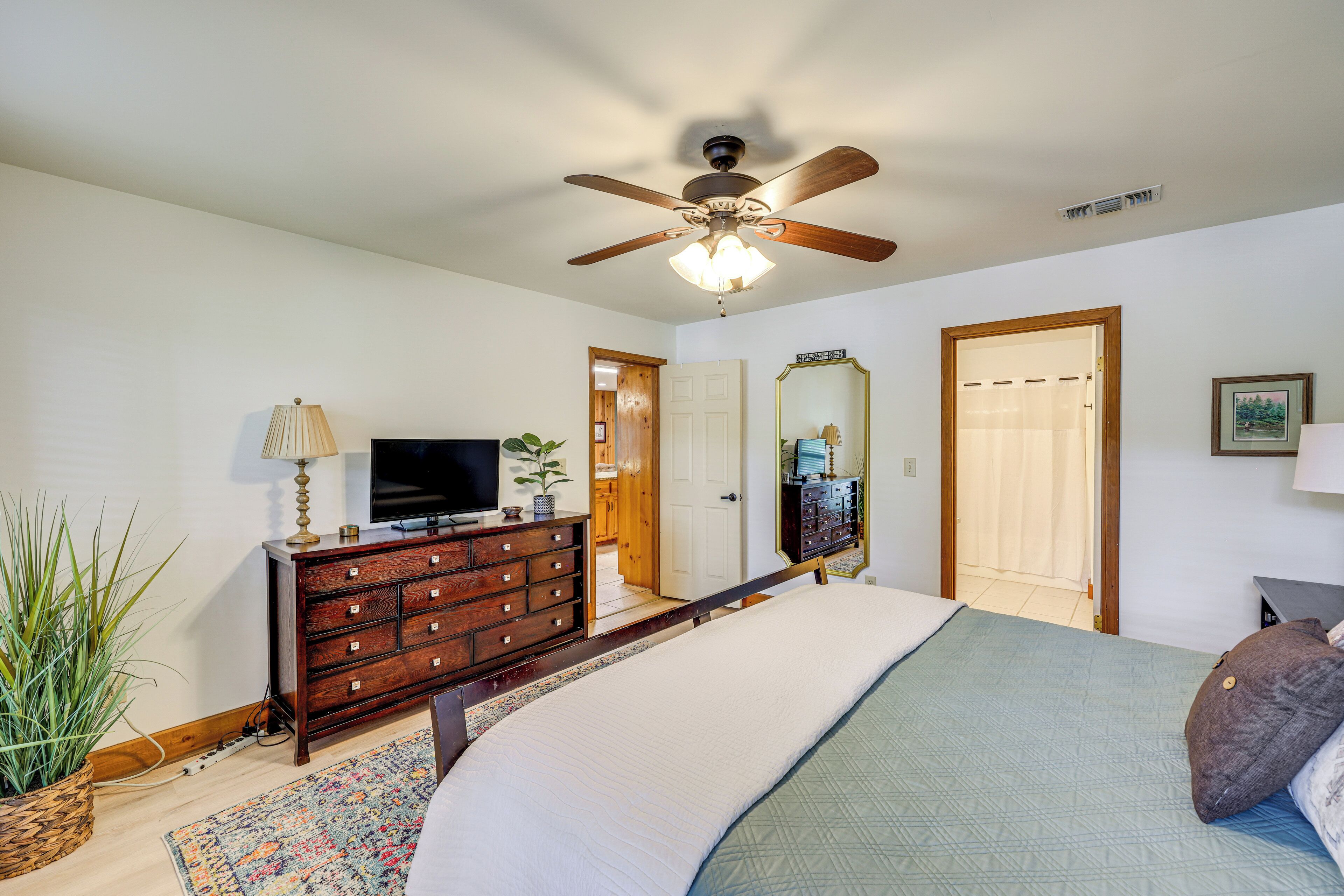 A bedroom with a bed, dresser, television, ceiling fan, and a door leading to another room.