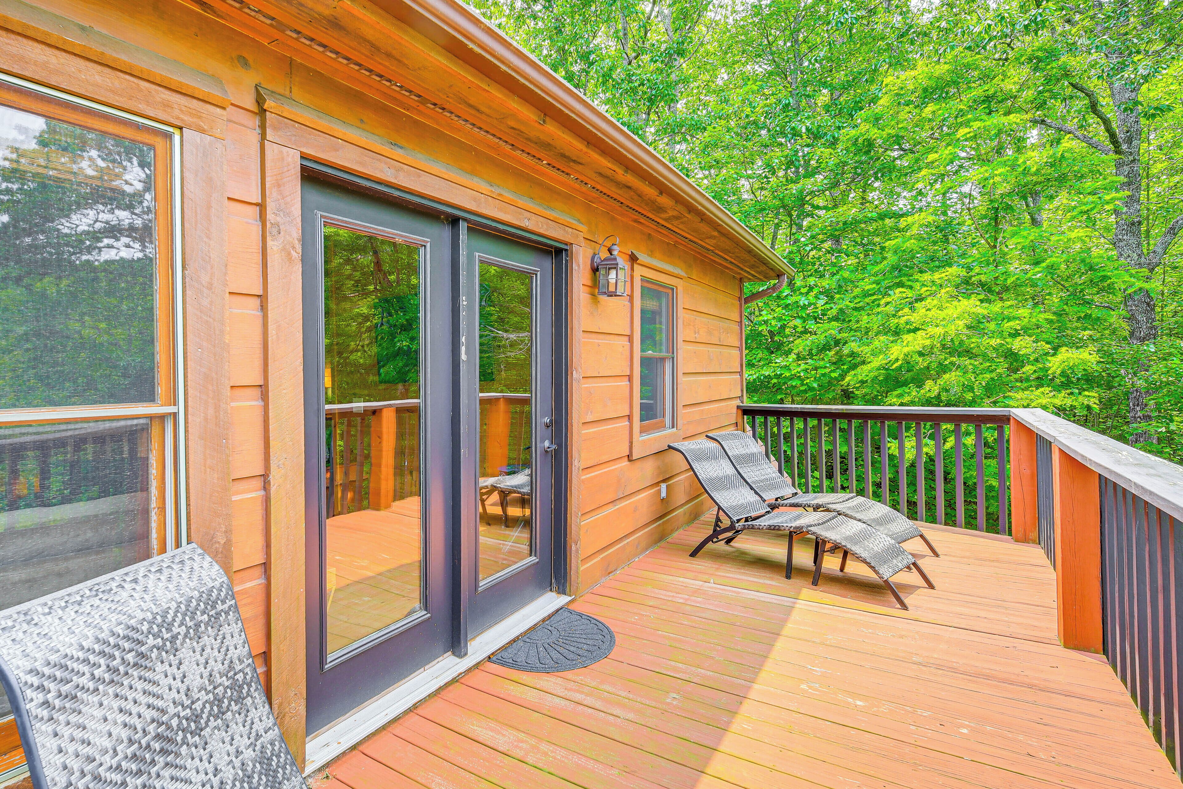A wooden deck with two lounge chairs and a table, a sliding glass door, and a view of a forested area.