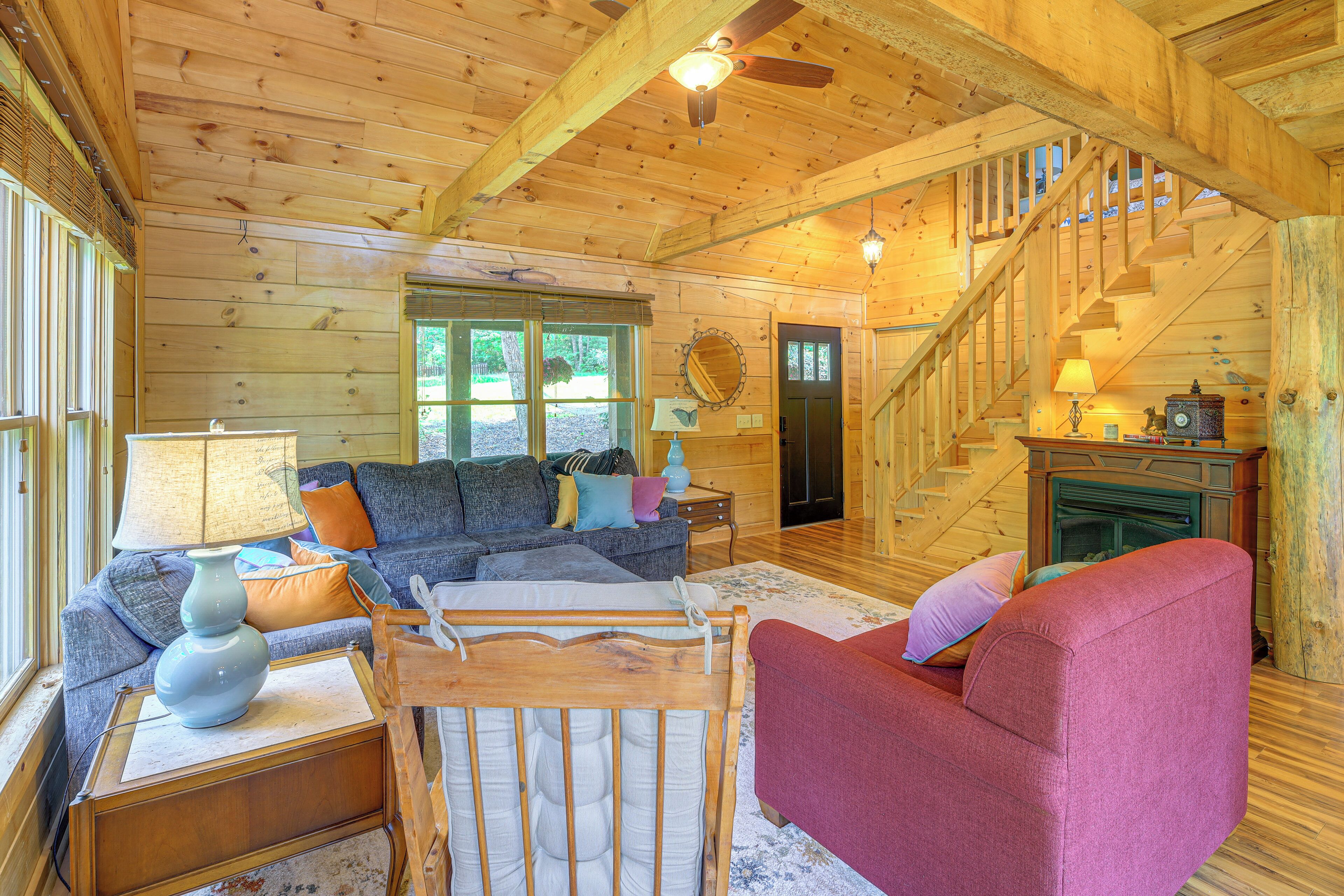 A cozy cabin interior with a wooden ceiling, a staircase, a living room with a sofa and armchairs, and a fireplace.