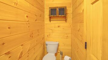 Une petite salle de bain aux murs lambrissés, équipée de toilettes, d’une fenêtre et d’un rouleau de papier toilette.