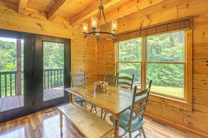A wooden cabin interior with a dining table, chairs, a chandelier, and large windows offering a view of the forest. - Blue Ridge Mtns Retreat w/ Views, 1 Mi to Parkway! (Hillsville)