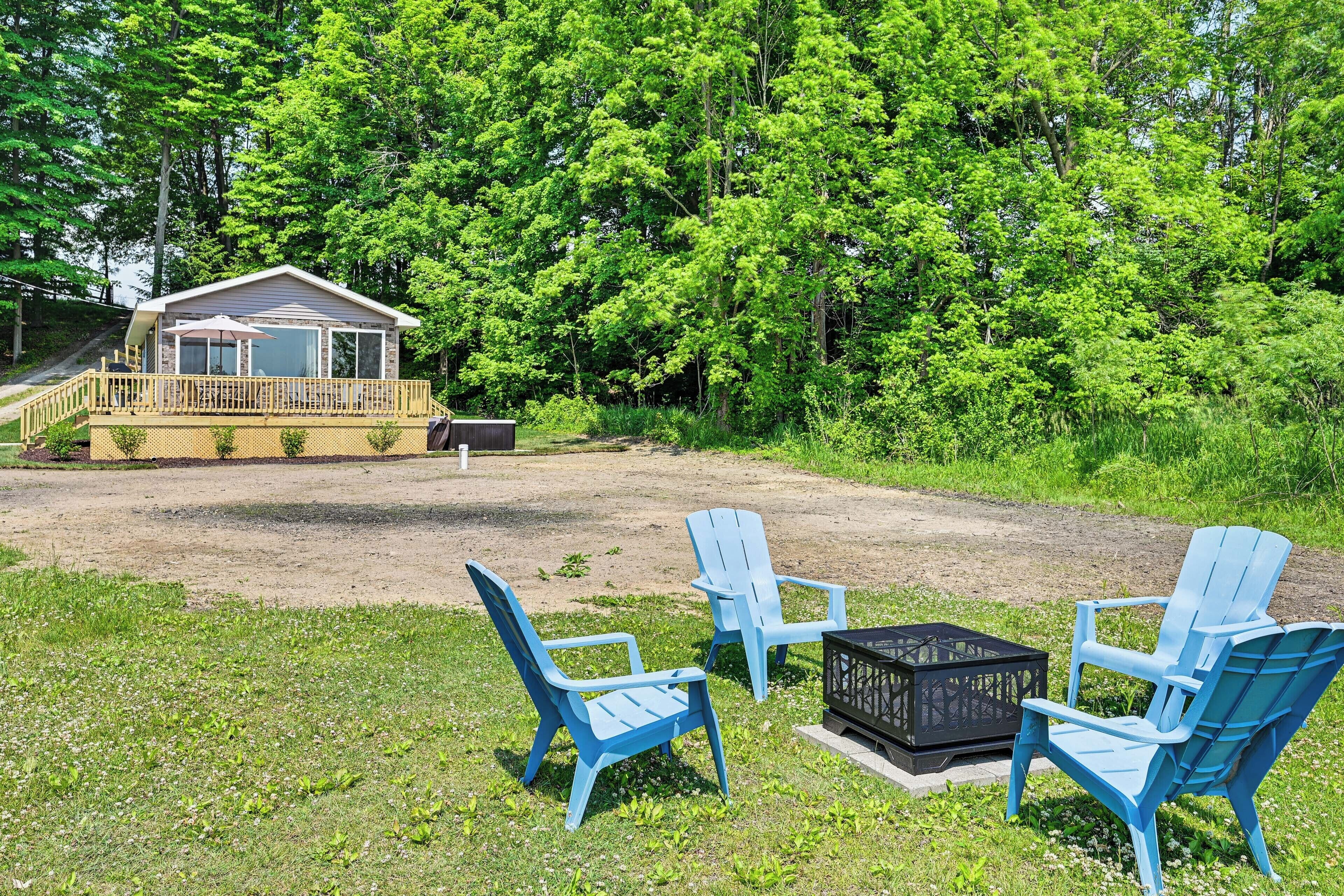 A cabin with a deck, surrounded by dense forest. There are three Adirondack chairs and a black lantern on the deck.