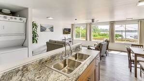 Une cuisine moderne dotée d’un plan de travail en granit, d’un évier en acier inoxydable et d’un four encastré. On y trouve un coin repas avec une table et des chaises en bois, ainsi qu’une grande fenêtre offrant une vue sur un plan d’eau et la verdure environnante.
