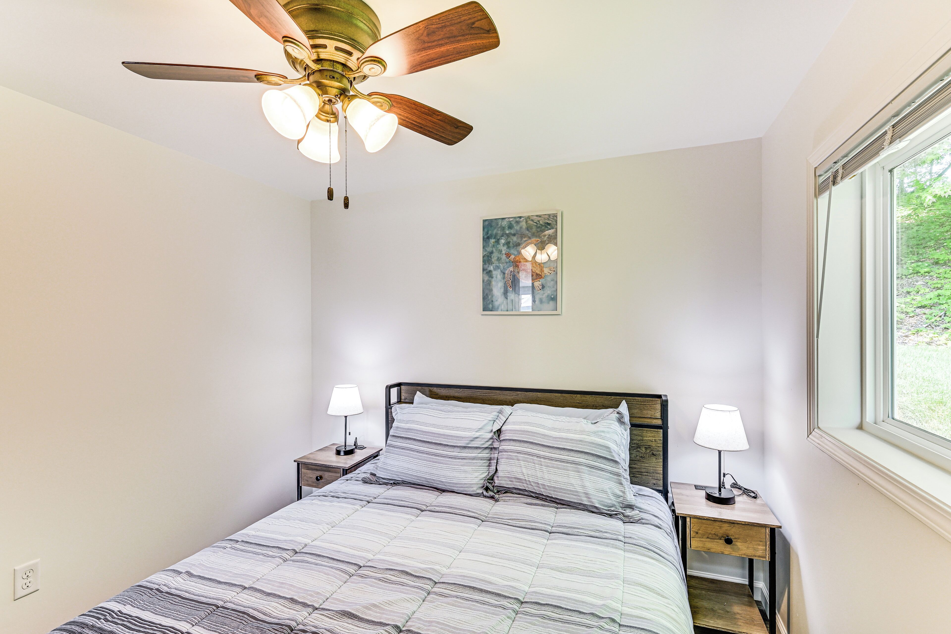 A bedroom with a bed, two bedside tables with lamps, a ceiling fan, a window, and a framed picture on the wall.